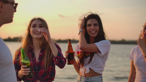 Two-female-students-are-touching-their-long-hair-and-talking-with-a-friend-on-the-beach-party-with-beer.-A-great-summer-open-air-party-next-to-the-lake-at-sunset.